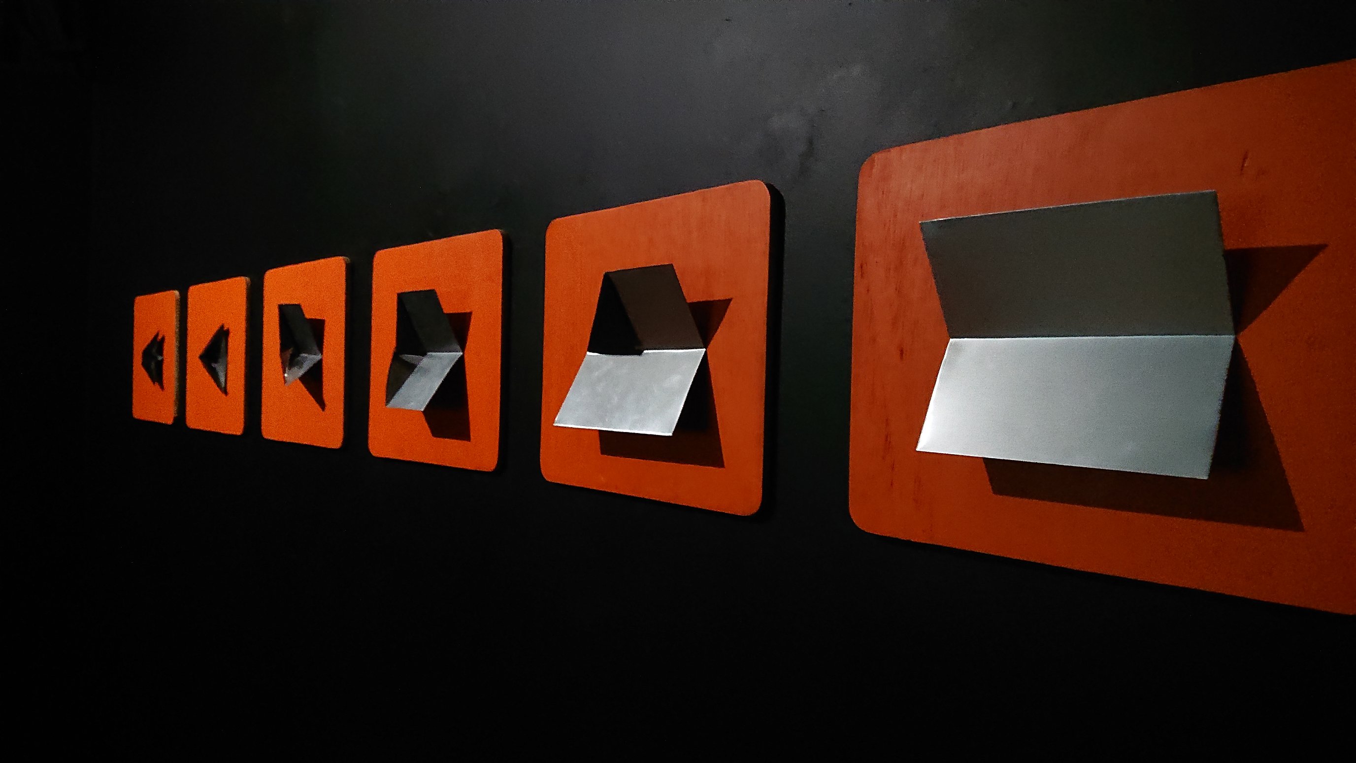 Six rectangular sheets of wood, painted orange, are mounted in a line across a black wall. Each sheet has one sheet of folded aluminium mounted on it, showing a progression of folds that you'd use to turn a rectangular piece of paper into a paper aeroplane
