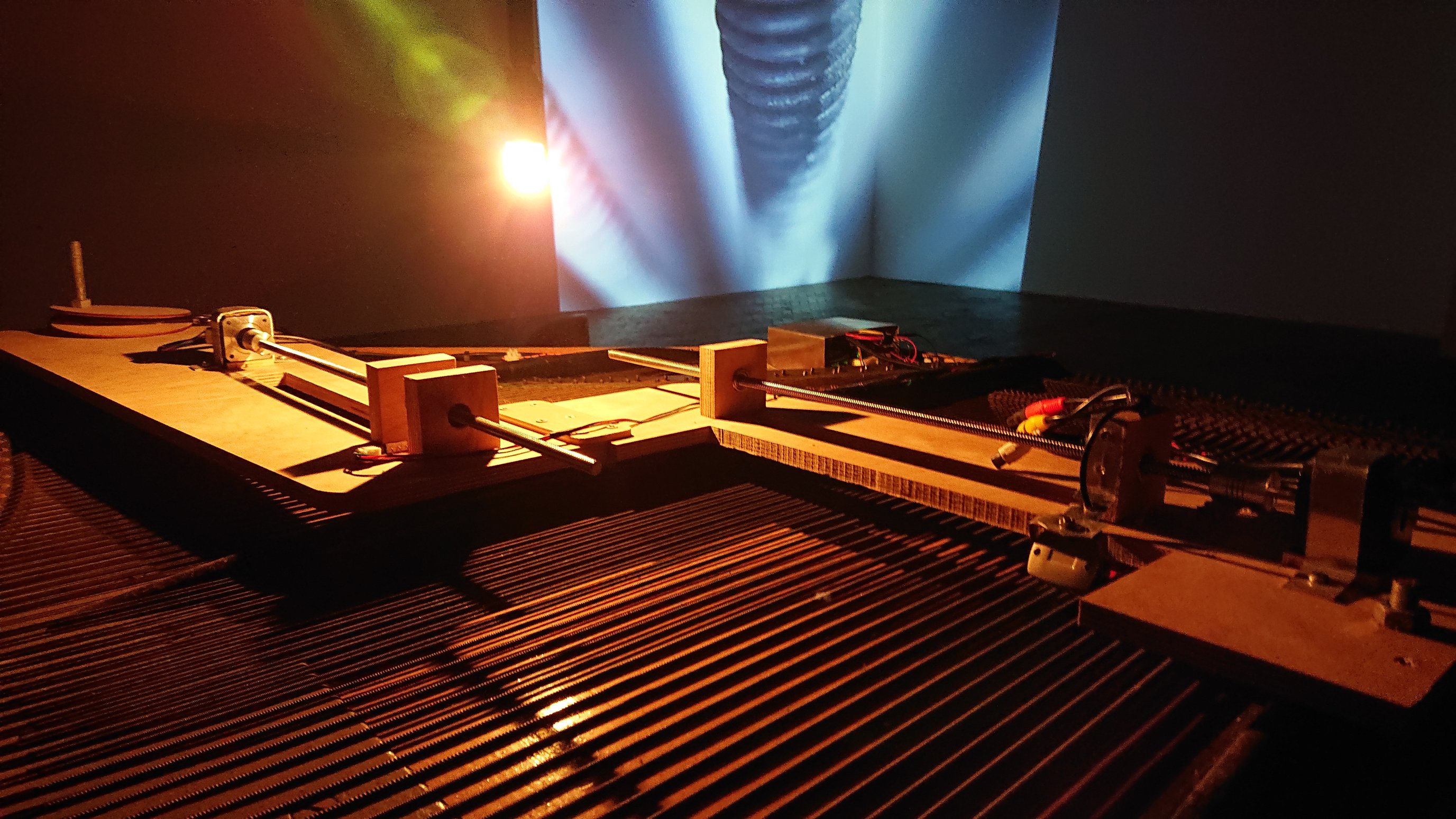 A view of the Tide Piano: the frame of strings mounted horizontally with a plywood bridge running across, perpendicular to the strings. The bridge is split into two halves: each of which has a stepper motor and leadscrew mounted on top, driving the plectrum across the strings. In the background of the shot we can see the rest of the gallery with the video feed showing the close-up coil of a piano string projected onto the corner wall